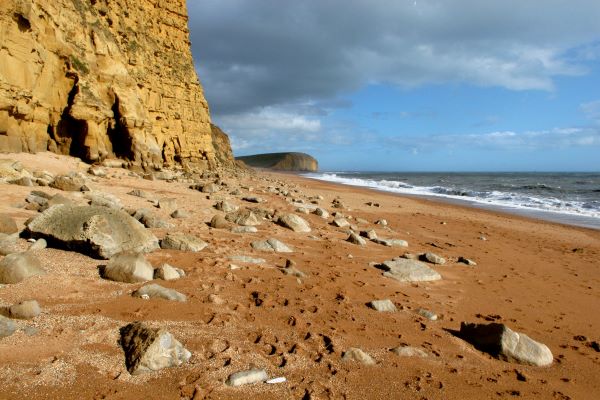 A close up view of the golden sandy-coloured cliffs of West Bay. The sky behind is grey and stormy. There are some large light sandy-coloured boulders on the sandy/tiny pebble beach. 